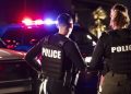 Rear view of two multiracial police officers standing side by side in front of their police vehicles with emergency lights on. They are working at night in a city. The policewoman is mixed race, African-American, Asian and Hispanic, in her 40s. Her partner is a young Hispanic man in his 20s.