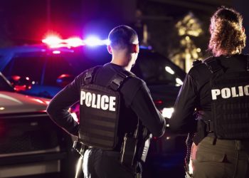 Rear view of two multiracial police officers standing side by side in front of their police vehicles with emergency lights on. They are working at night in a city. The policewoman is mixed race, African-American, Asian and Hispanic, in her 40s. Her partner is a young Hispanic man in his 20s.