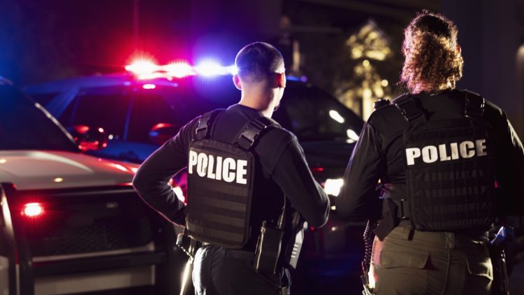 Rear view of two multiracial police officers standing side by side in front of their police vehicles with emergency lights on. They are working at night in a city. The policewoman is mixed race, African-American, Asian and Hispanic, in her 40s. Her partner is a young Hispanic man in his 20s.