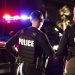Rear view of two multiracial police officers standing side by side in front of their police vehicles with emergency lights on. They are working at night in a city. The policewoman is mixed race, African-American, Asian and Hispanic, in her 40s. Her partner is a young Hispanic man in his 20s.
