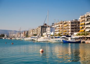 Beautiful promenade in Volos, Greece, by the Aegean sea's Pagasetic Gulf. The Pelion mountain range, port, boats, apartment buildings and restaurants are visible in the image and so are some unrecognizable people walking.