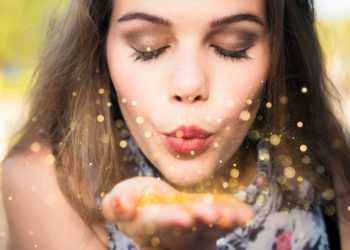 A young woman closing her eyes as she blows golden confetti from her hand, and makes a wish.