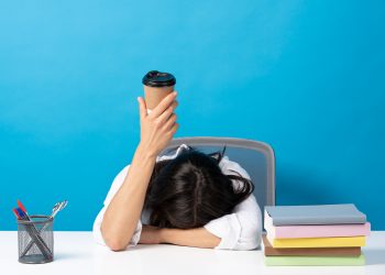 Woman sleeping head on desk showing disposable cup of coffee isolated on blue background. Caffeine addiction concept (iStock)
