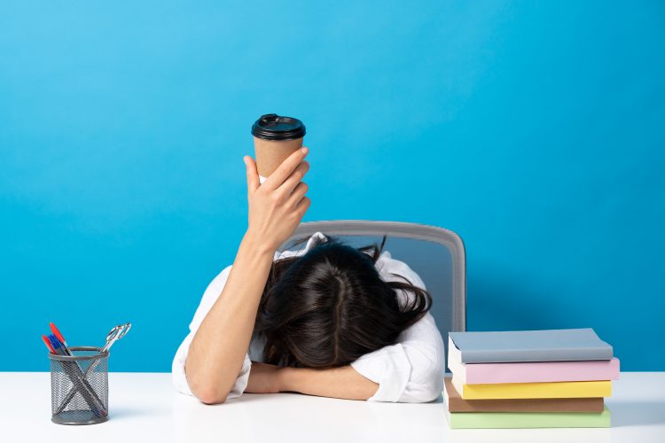 Woman sleeping head on desk showing disposable cup of coffee isolated on blue background. Caffeine addiction concept (iStock)