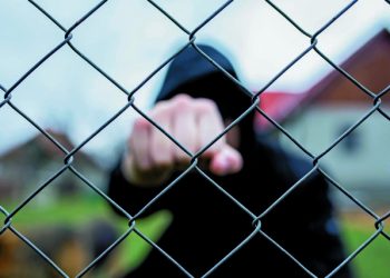 Aggressive teenage boy showing hes fist behind wired fence at the correctional institute, focus on the wired fence, conceptual image of juvenile delinquency .