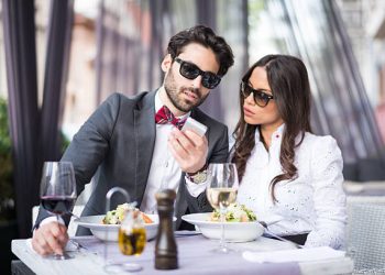 Couple of young people wearing sunglasses having a meal in restaurant. Man is showing mobile phone content to his female friend.