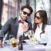 Couple of young people wearing sunglasses having a meal in restaurant. Man is showing mobile phone content to his female friend.