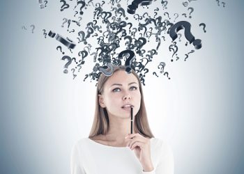 Attractive young woman with fair hair wearing white is holding a pen near her full lips, looking up and thinking. A gray wall background with question marks