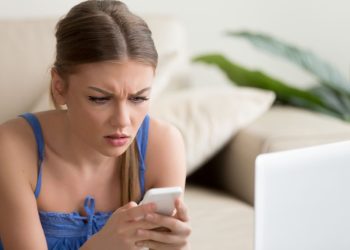 Worried young lady lying on sofa in front of laptop and looking on cellphone in her hands with confusion. Stressed lady reading unexpected message with bad news, received sad e-mail on mobile phone