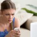 Worried young lady lying on sofa in front of laptop and looking on cellphone in her hands with confusion. Stressed lady reading unexpected message with bad news, received sad e-mail on mobile phone